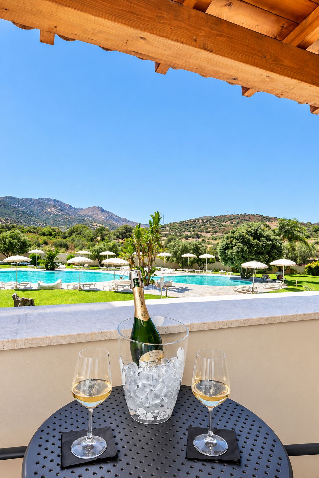 A table with a bottle of wine and two glasses, overlooking a pool and a mountainous landscape.