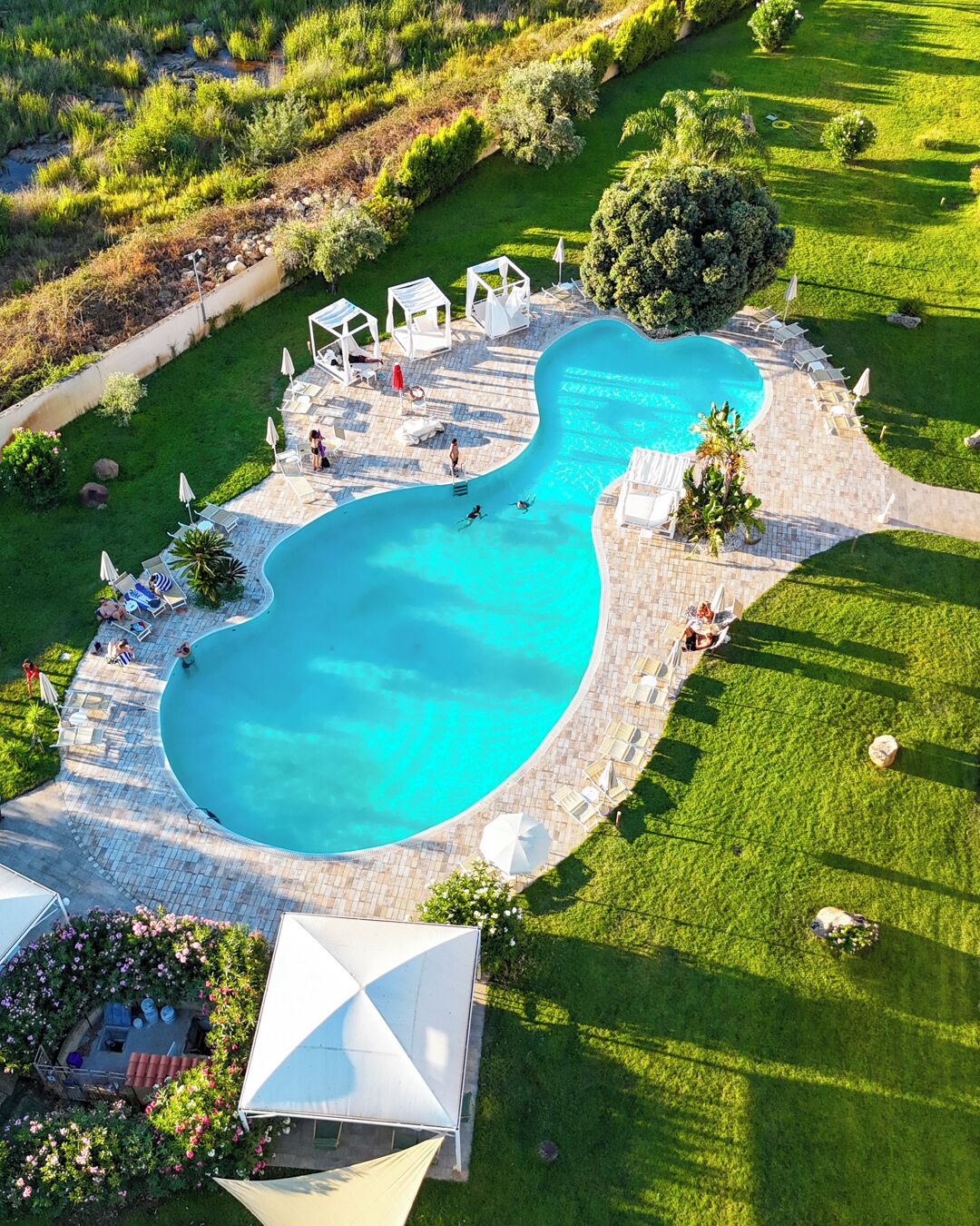 Luftaufnahme eines Swimmingpools mit umlaufender Terrasse, inklusive Liegestühlen und Sonnenschirmen.