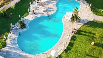Luftaufnahme eines Swimmingpools mit umlaufender Terrasse, inklusive Liegestühlen und Sonnenschirmen.