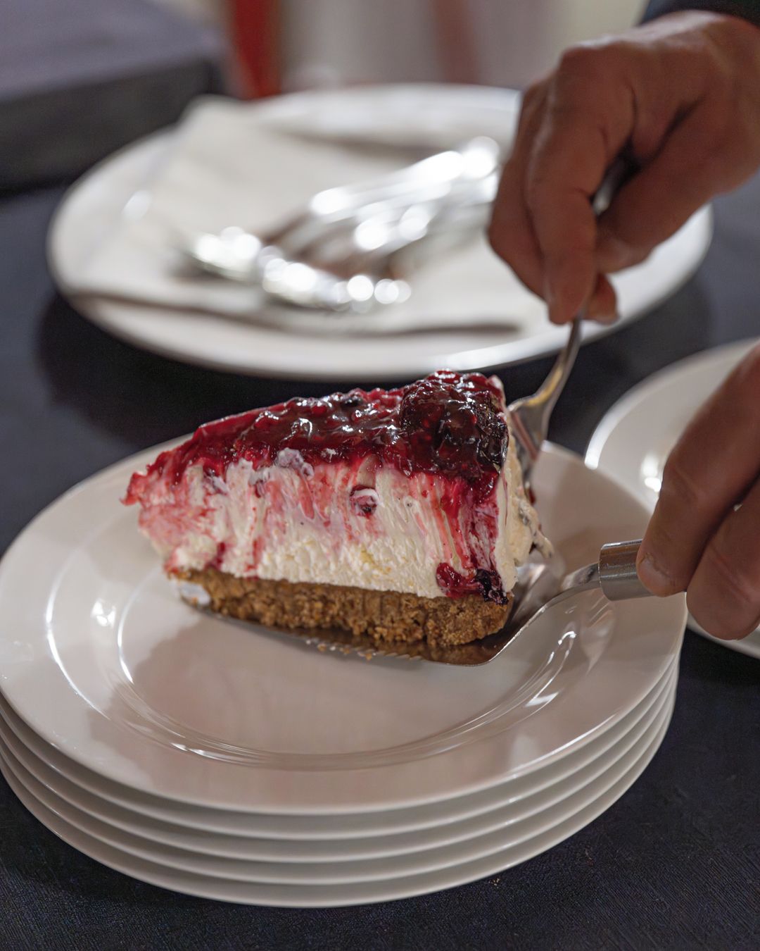 Una porción de pastel de queso en un plato blanco con un tenedor, sobre una mesa con platos apilados.