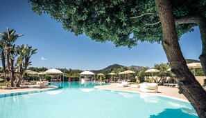 Una piscina con sillas de descanso y sombrillas, rodeada de árboles y una montaña al fondo.