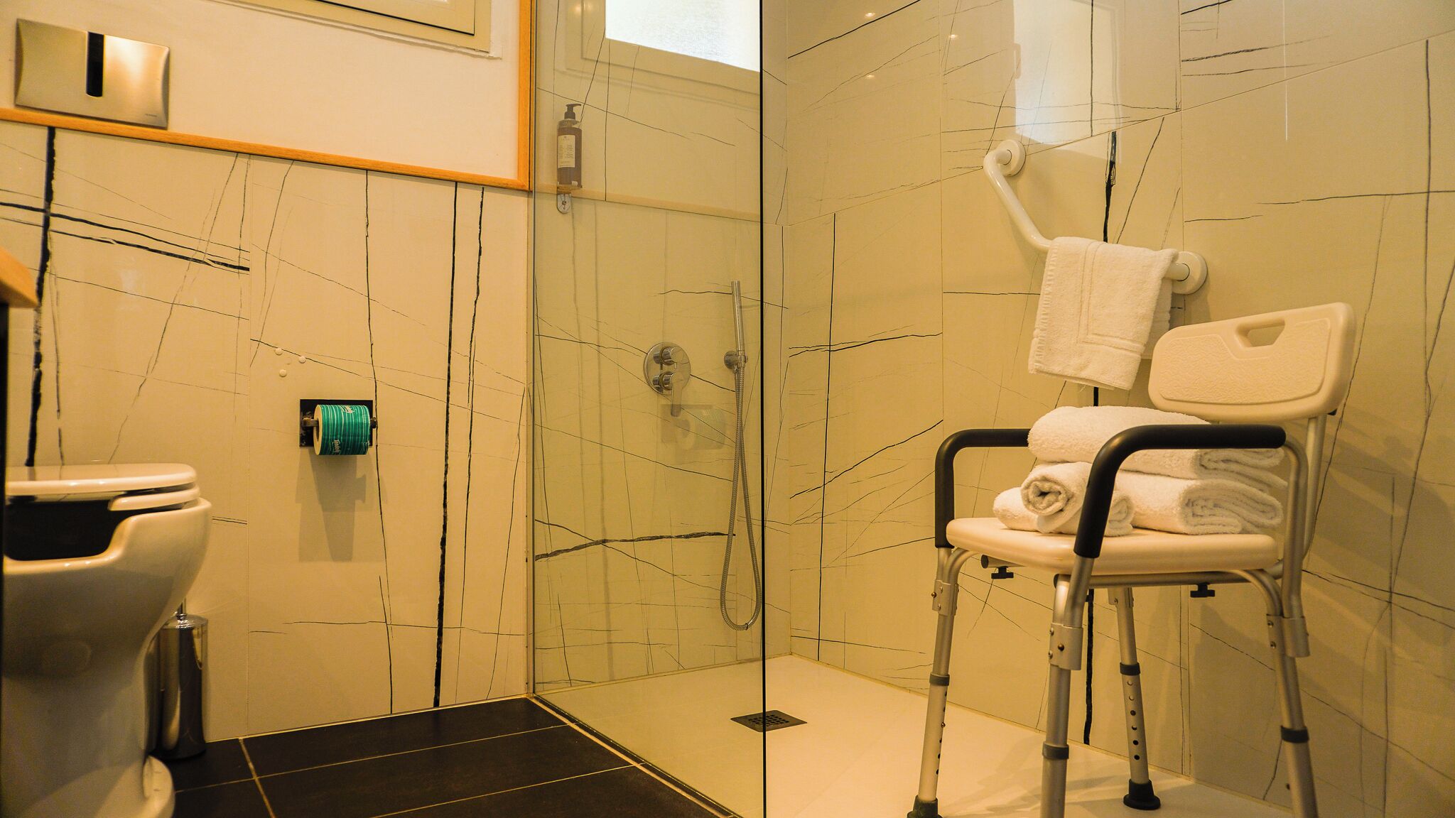 Baño con cabina de ducha de vidrio, una silla de ruedas y un perchero para toallas.