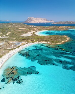 Vista aérea de una zona costera con playa, aguas turquesas cristalinas y montañas al fondo.