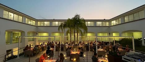 Un espacio al aire libre para comer con varias mesas y sillas, rodeado por un edificio blanco con ventanales amplios.
