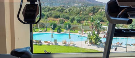 Ein Heimfitnessraum mit Trainingsgeräten, Blick auf einen Swimmingpool und eine Berglandschaft.