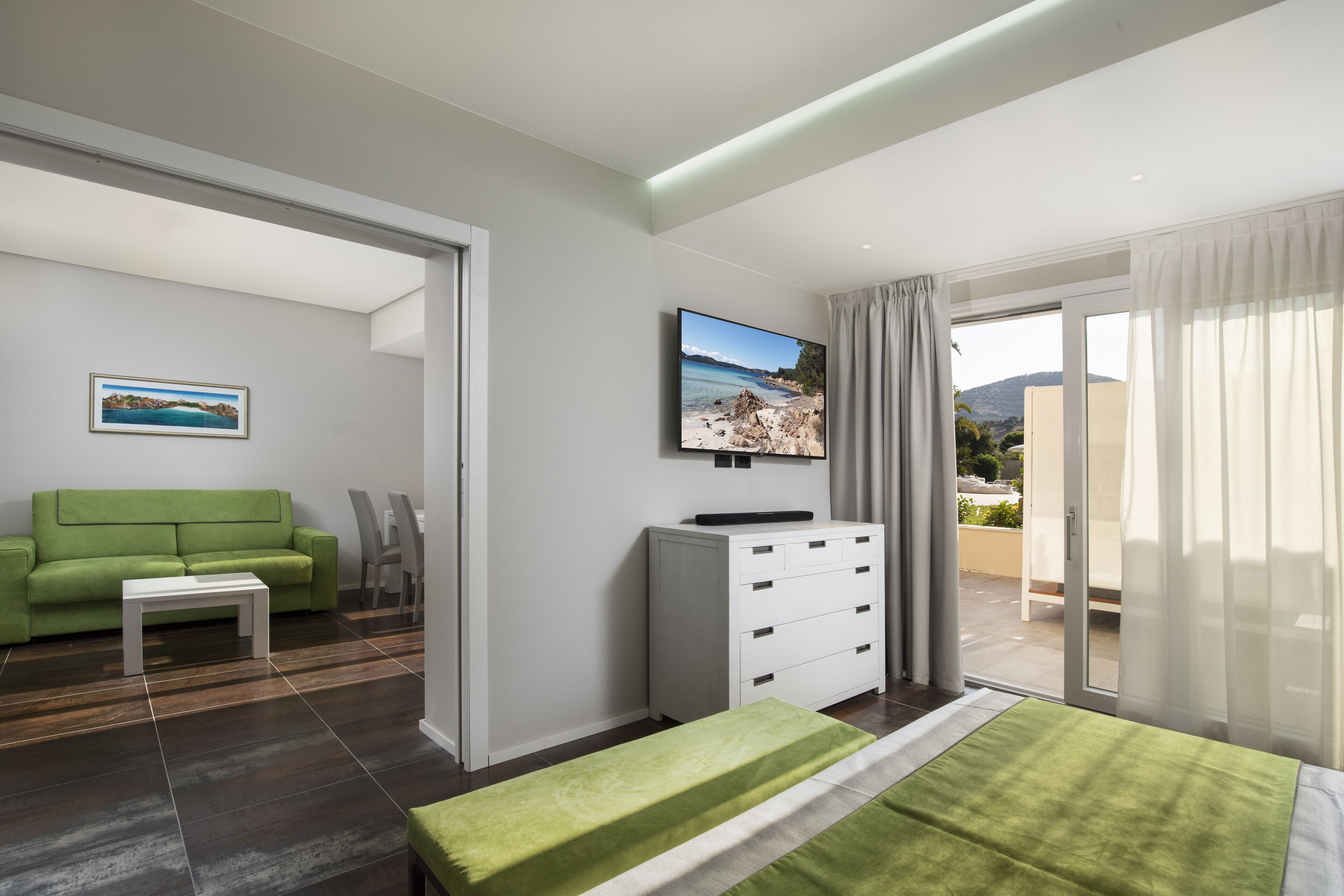 A modern hotel room with a green sofa, a white coffee table, a flat-screen TV mounted on the wall, a white dresser, and a balcony with a view of the beach.