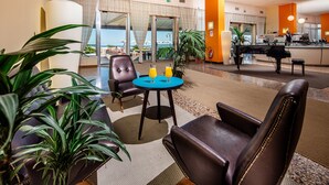 Una hall d'hotel con poltrone in pelle, un pianoforte e vista sulla spiaggia attraverso ampie finestre.