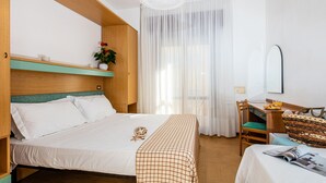 A hotel room with a bed, a desk, a chair, a window with sheer curtains, and a hanging light fixture. - Hotel Astor (Jesolo)