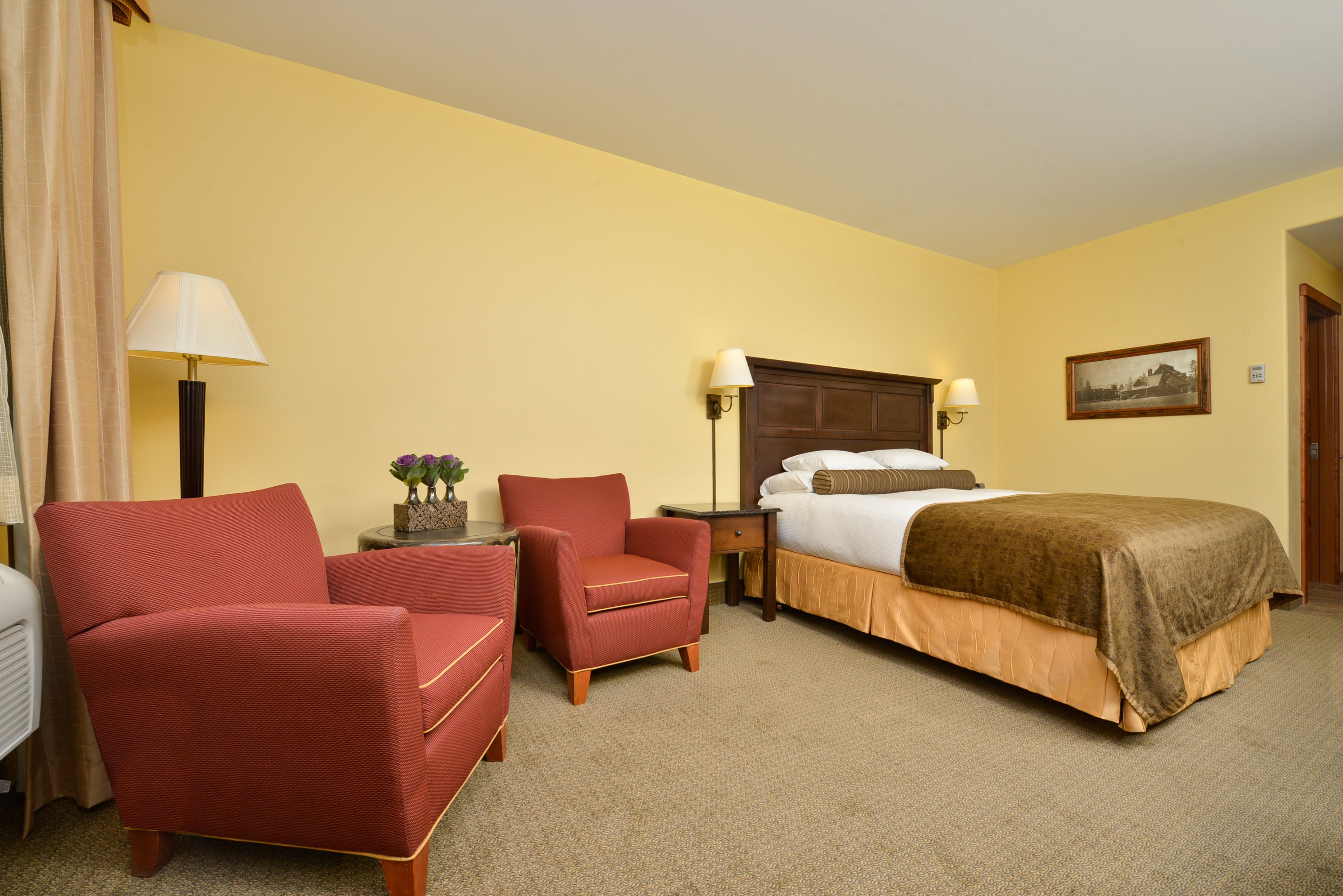 A hotel room with a bed, two chairs, a nightstand, and a lamp.