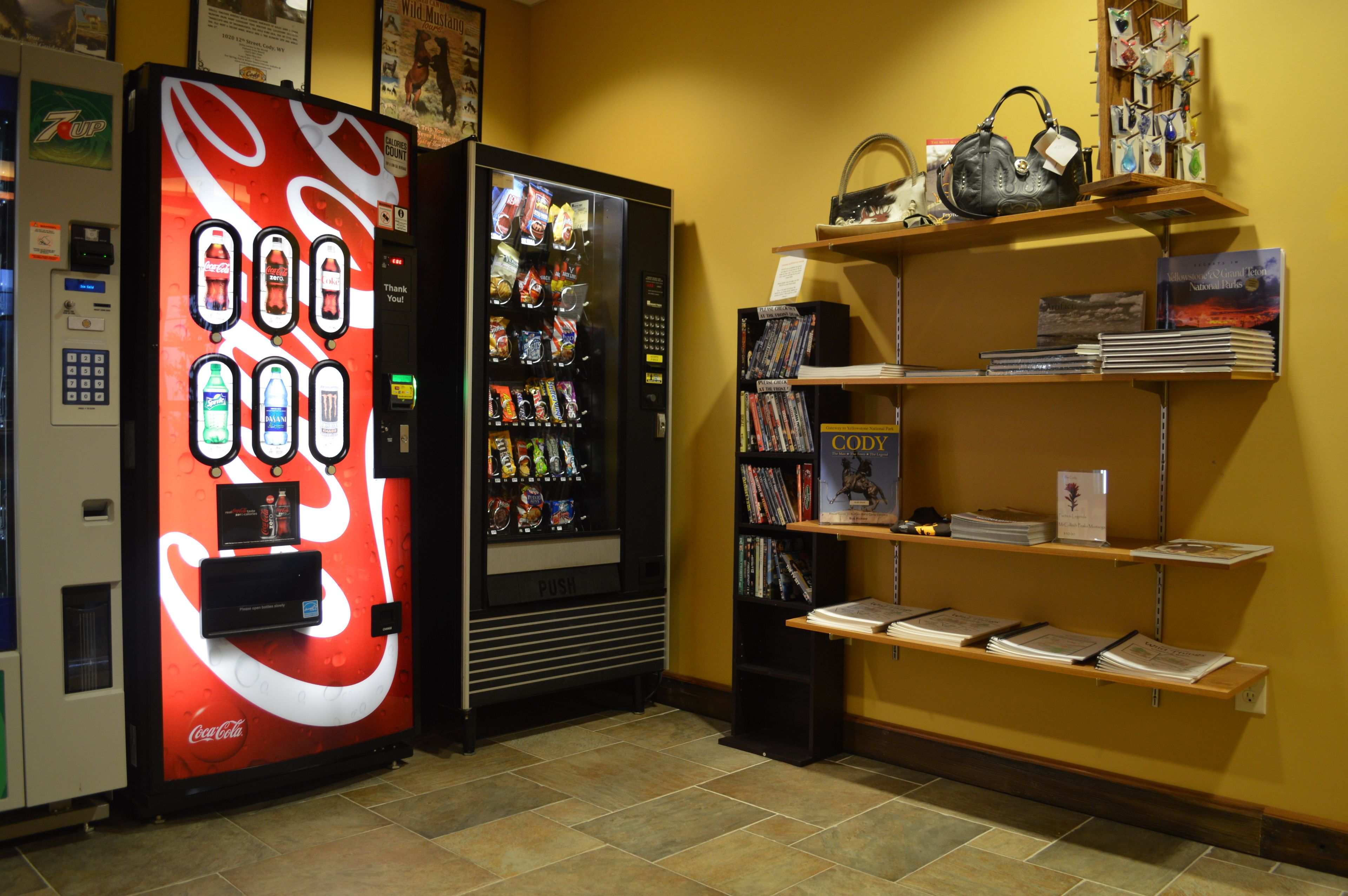 A vending machine with a Coca-Cola logo, a snack vending machine, and a bookshelf with books and a handbag.