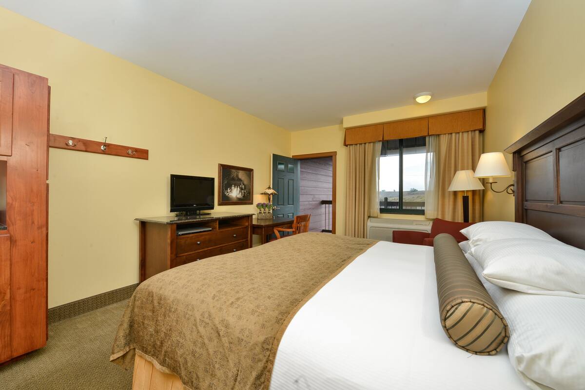 A hotel room with a bed, a desk with a television, a chair, a window with curtains, and a door.