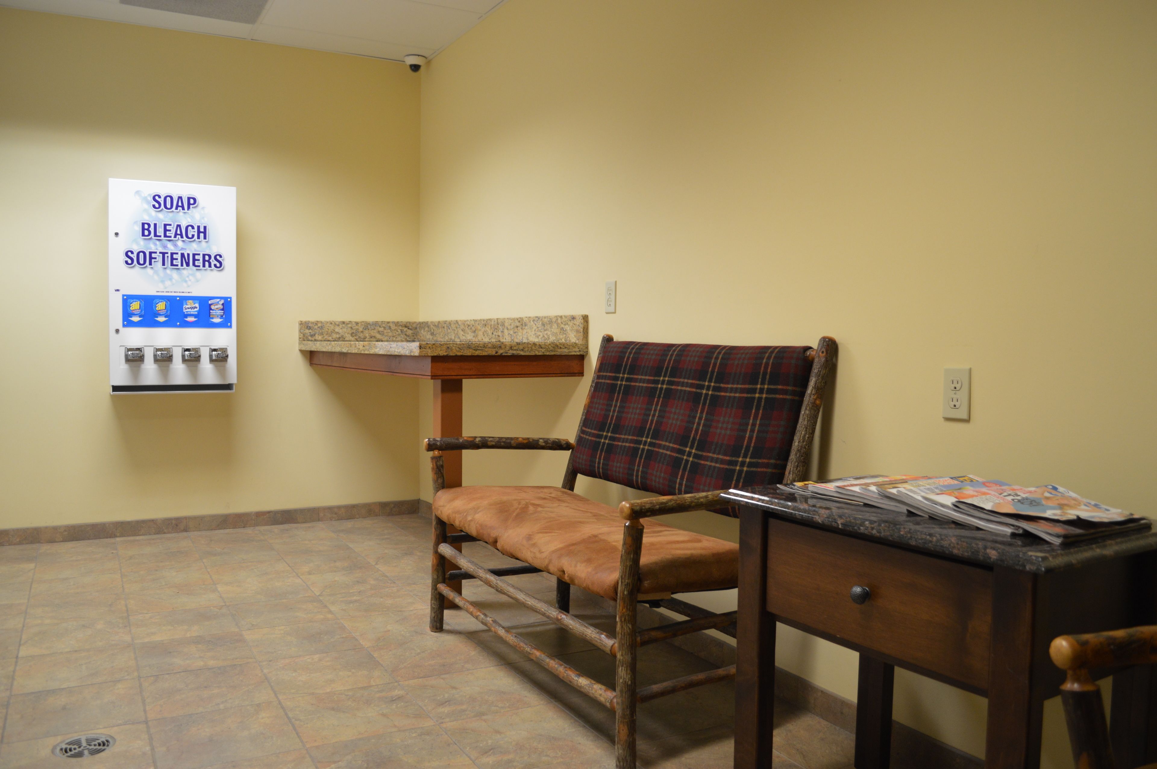 A room with a wooden chair, a small table, and a wall-mounted soap and bleach dispenser.