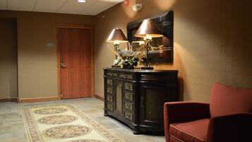 A hotel lobby with a wooden door, a dark-colored console table with two lamps, a red chair, and a patterned rug.
