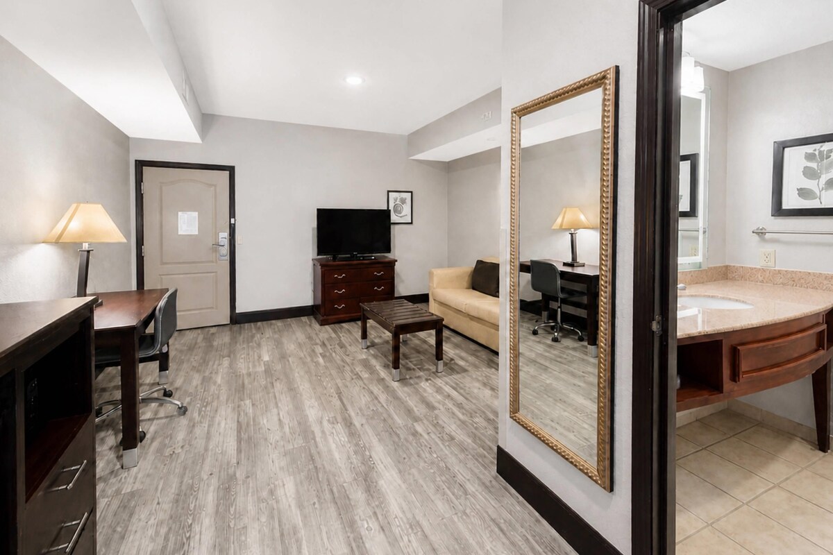 Een hotelkamer met een houten vloer, een bank, een bureau met stoel, een televisie, een dressoir, een grote spiegel en een badkamer met wastafel en toilet.