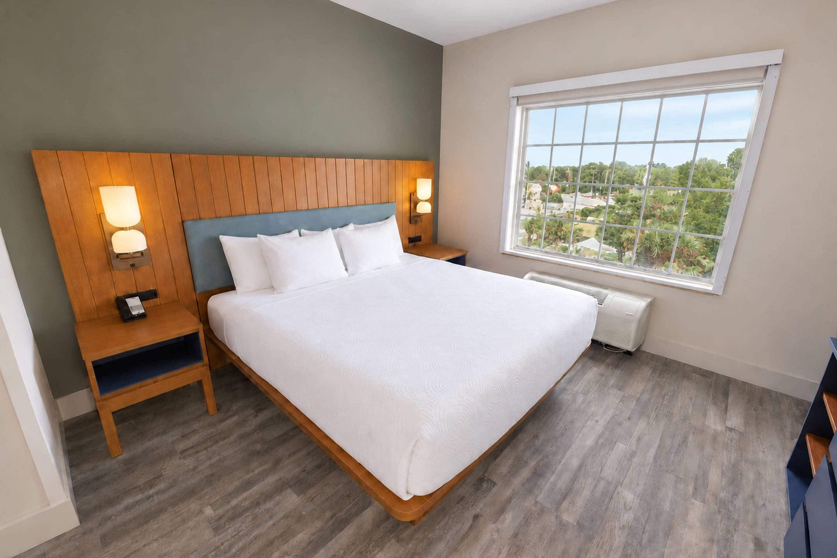 A hotel room with a large bed, wooden headboard, two bedside tables with lamps, a large window with a view, and a built-in air conditioning unit.