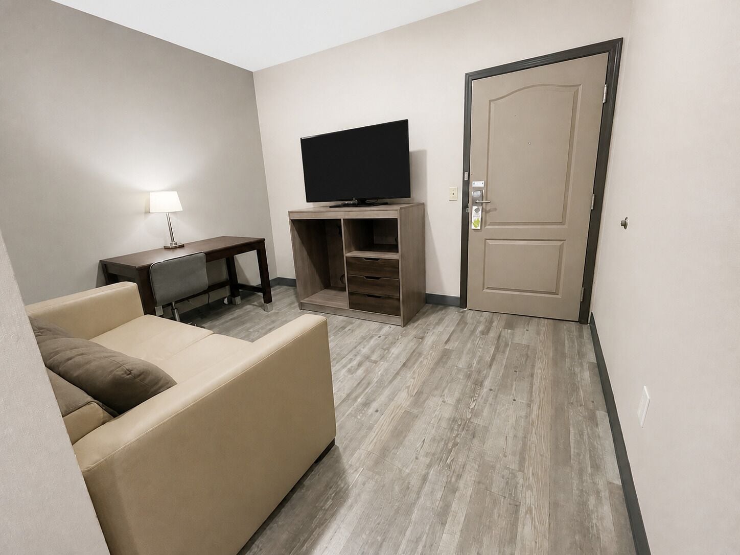 A hotel room with a flat-screen TV on a wooden stand, a sofa, a desk with a lamp, and a door.