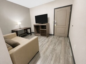Habitación de hotel con una televisión de pantalla plana sobre un soporte de madera, un sofá, un escritorio con lámpara y una puerta.