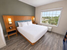 A hotel room with a large bed, wooden headboard, bedside lamps, a nightstand, a window with a view, and a built-in heater.