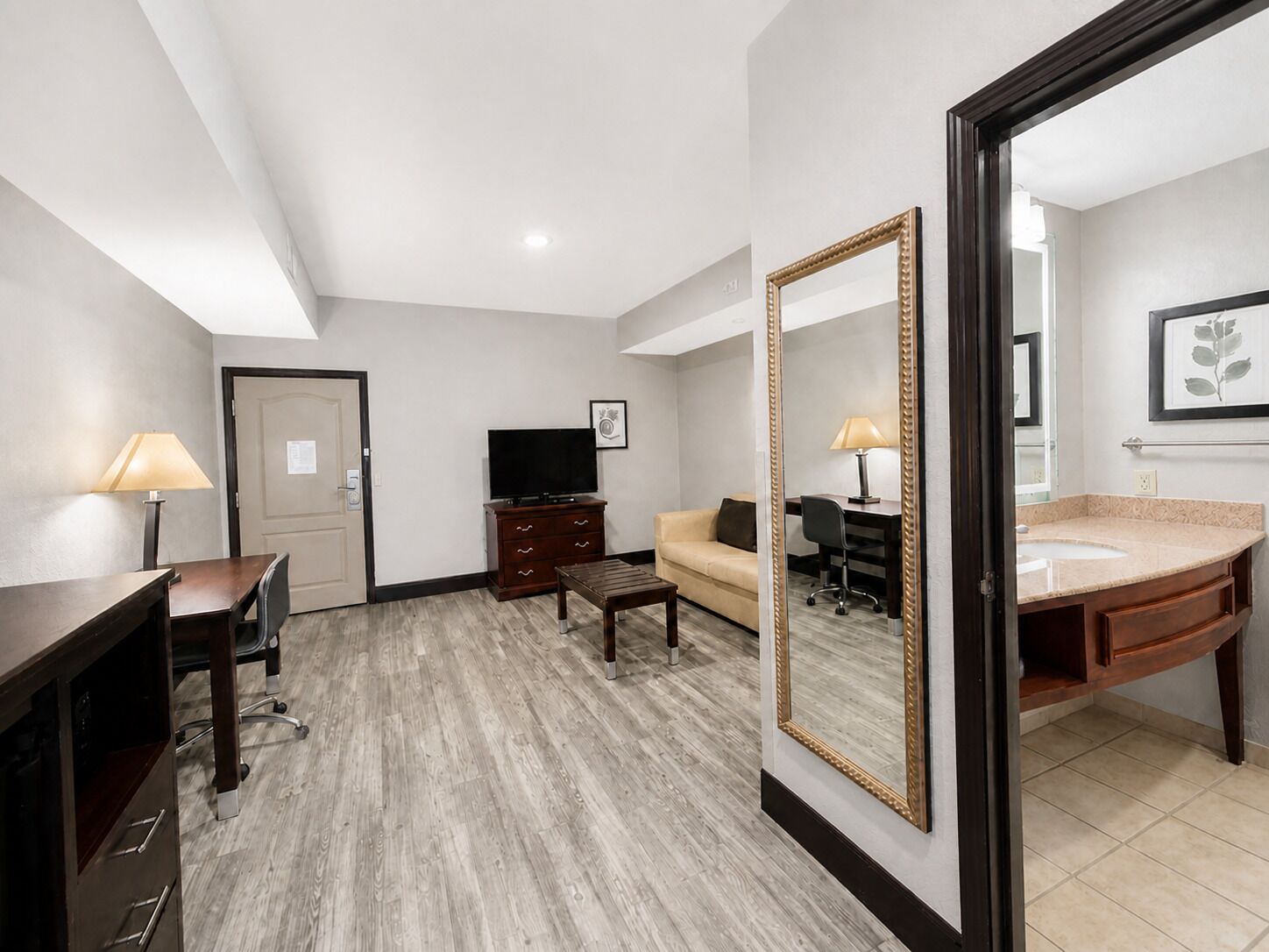A hotel room with a wooden desk, a television, a sofa, a coffee table, and a bathroom with a sink and mirror.