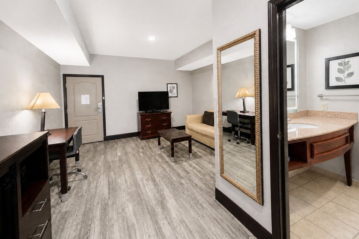 A hotel room with a wooden desk, a television, a sofa, a coffee table, and a bathroom with a sink and mirror.