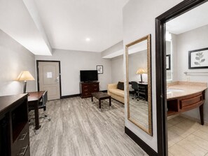 Habitación de hotel con escritorio de madera, televisor, sofá, mesa de centro y baño con lavamanos y espejo.