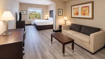 A hotel room with a bed, a sofa, a desk, and a coffee table. There are two lamps, a TV, and framed artwork on the walls.