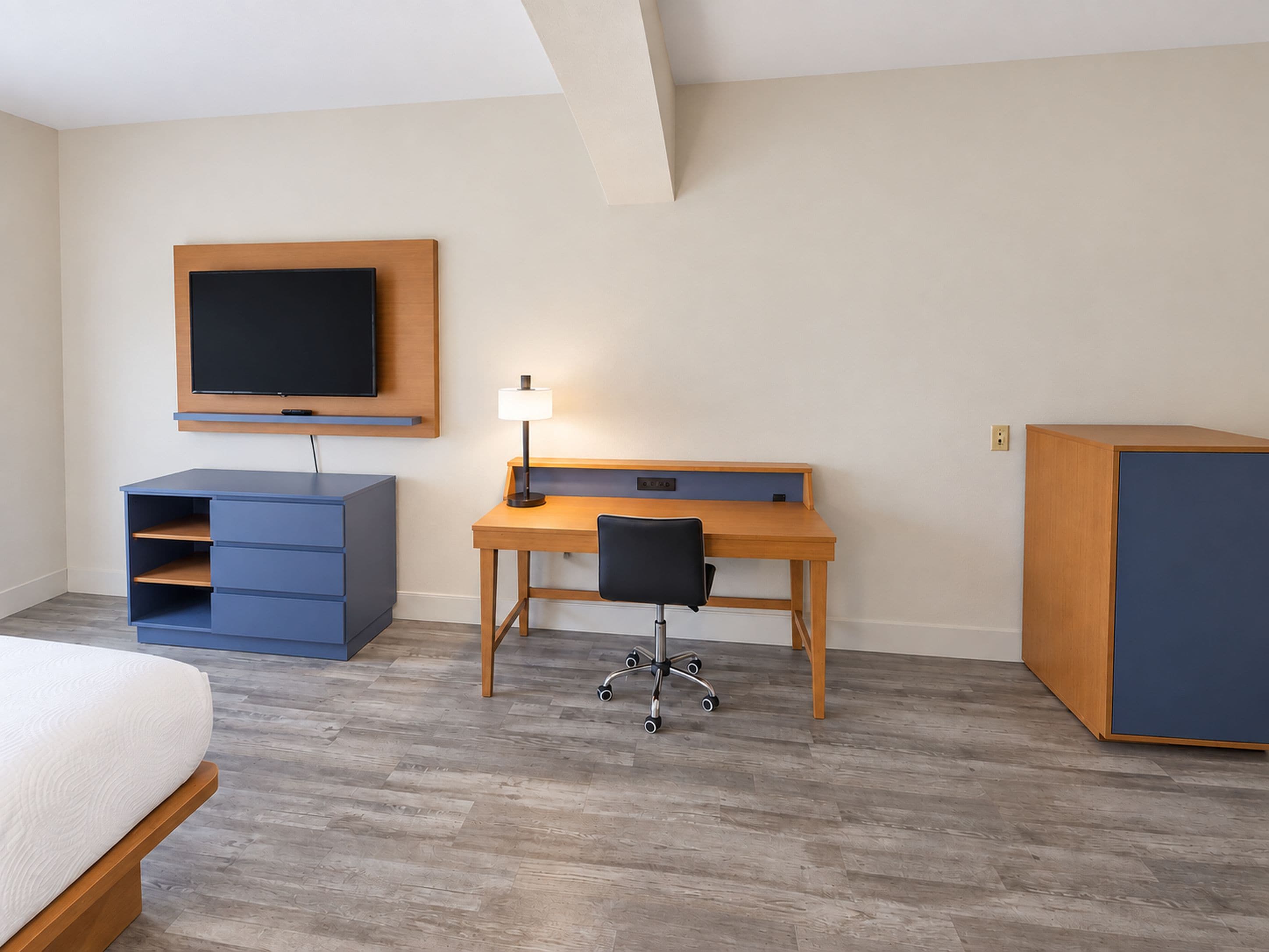 A hotel room with a bed, a desk with a chair, a television mounted on the wall, and a wardrobe.