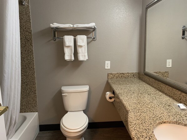 A bathroom with a toilet, a towel rack with two towels, a sink with a granite countertop, and a mirror. - Hawthorn Extended Stay by Wyndham Hobbs (Hobbs)