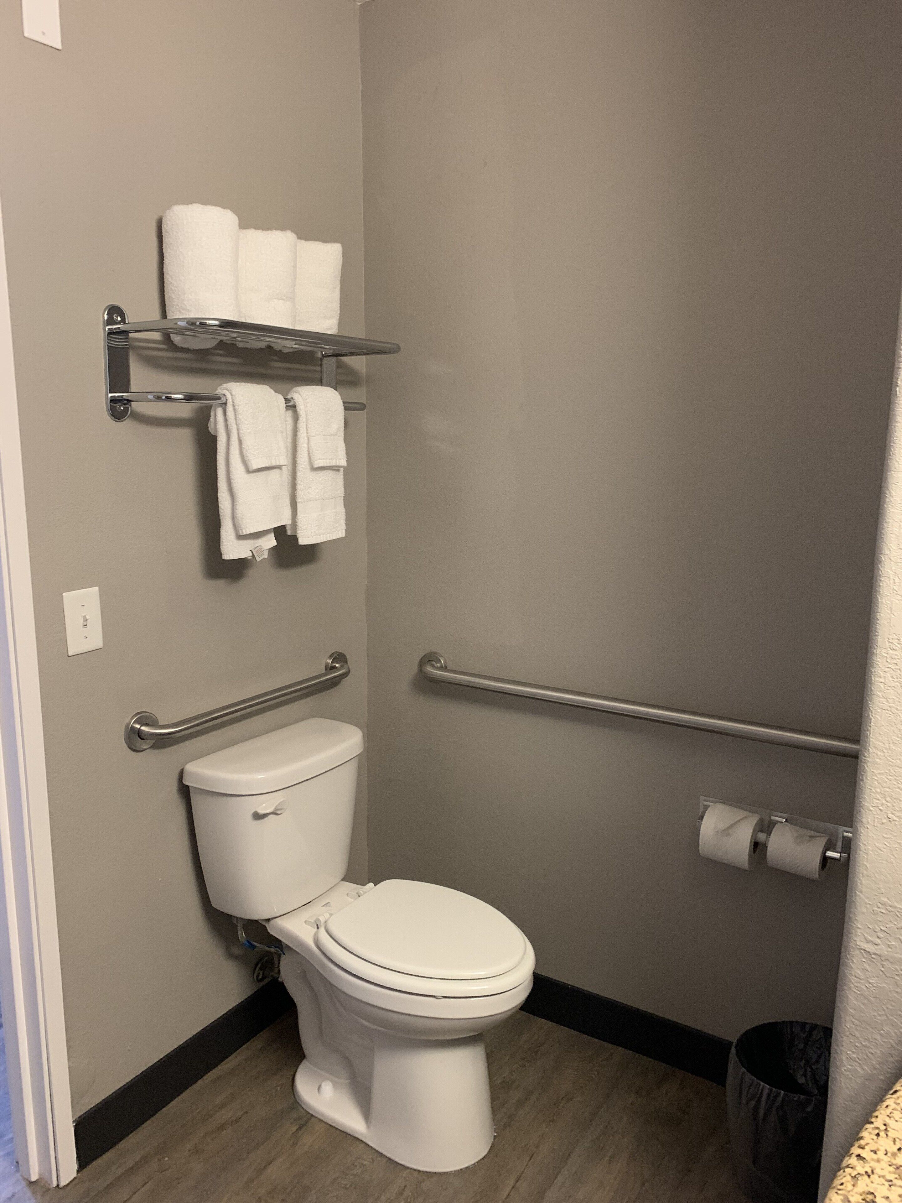 A bathroom with a white toilet, a handrail, a shelf with towels, and a trash can.