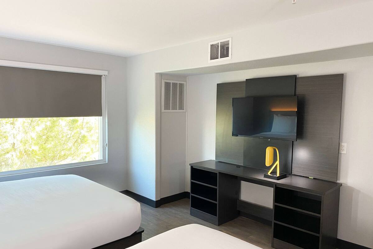 A modern hotel room with a bed, a desk with a TV, and a window with blinds.