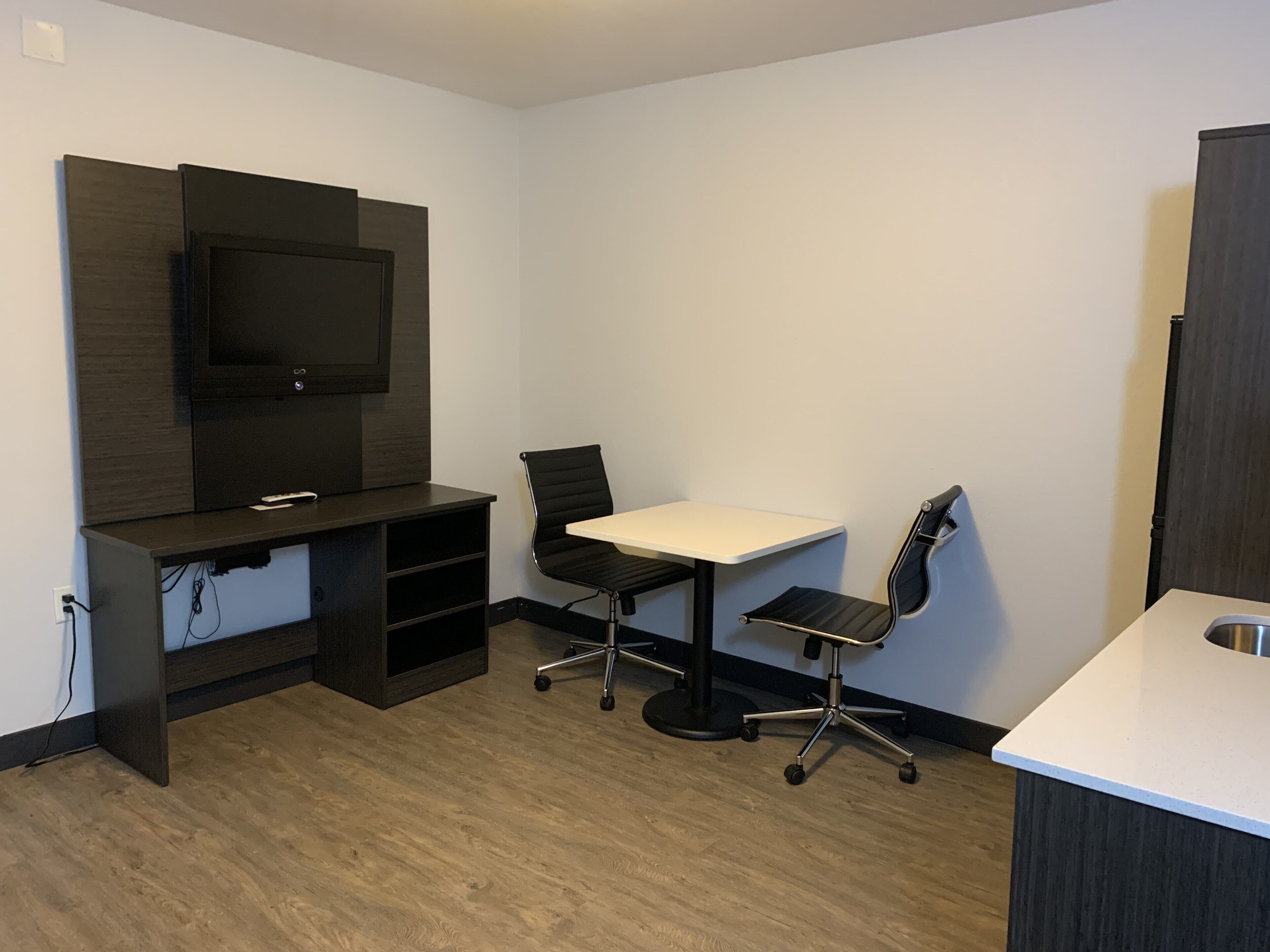 A small, modern room with a flat-screen TV mounted on the wall, a desk with a chair, and a kitchenette area.