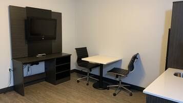 A small, modern room with a flat-screen TV mounted on the wall, a desk with a chair, and a kitchenette area.