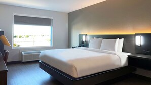 A modern hotel room with a large bed, a bedside table, a nightstand, a window with a view, and a wall-mounted light fixture.