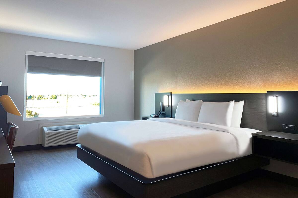 A modern hotel room with a large bed, a bedside table, a nightstand, a window with a view, and a wall-mounted light fixture.
