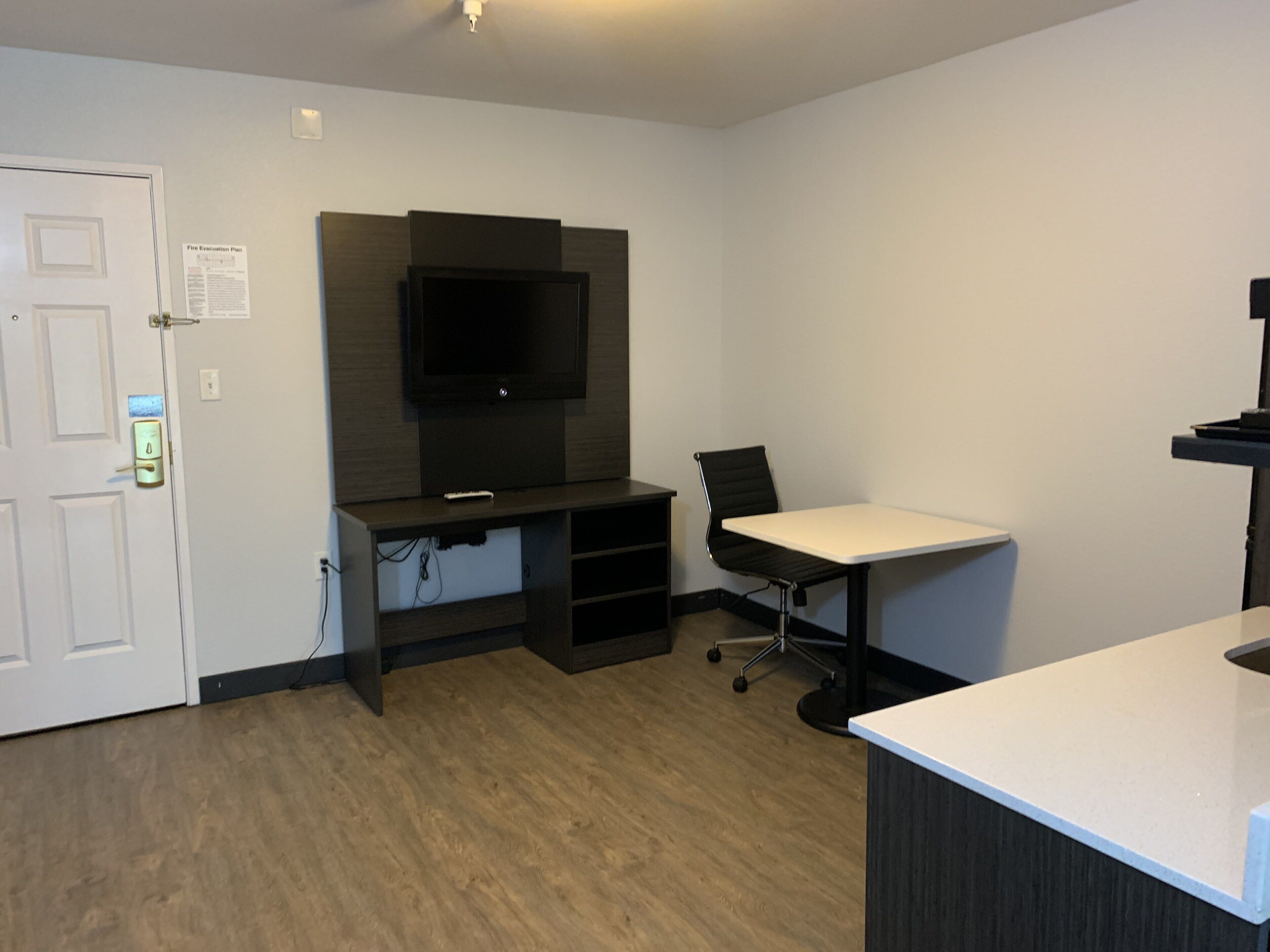 A room with a flat-screen TV mounted on a dark wooden wall, a white desk with a black office chair, and a white door with a lock.