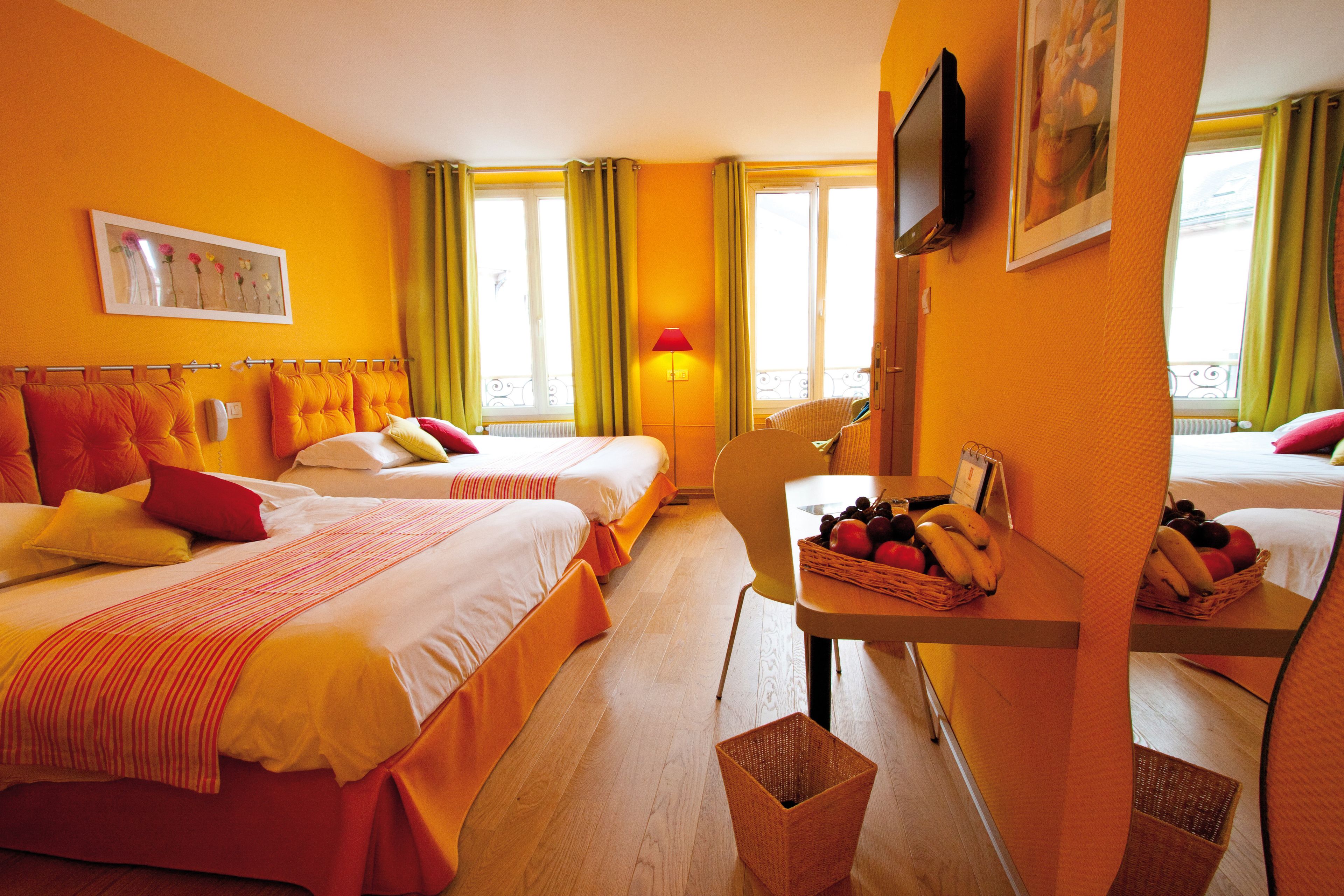 A hotel room with two beds, a desk with a basket of fruit, a lamp, and a TV on the wall.
