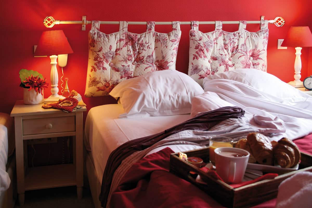 A hotel room with a bed, a nightstand, a lamp, a tray of breakfast items, and floral curtains.