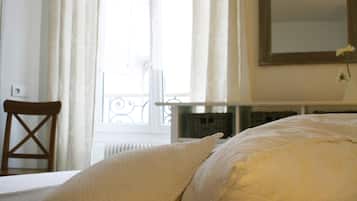 A bedroom with a bed, pillows, a chair, a window with curtains, and a mirror on the wall.