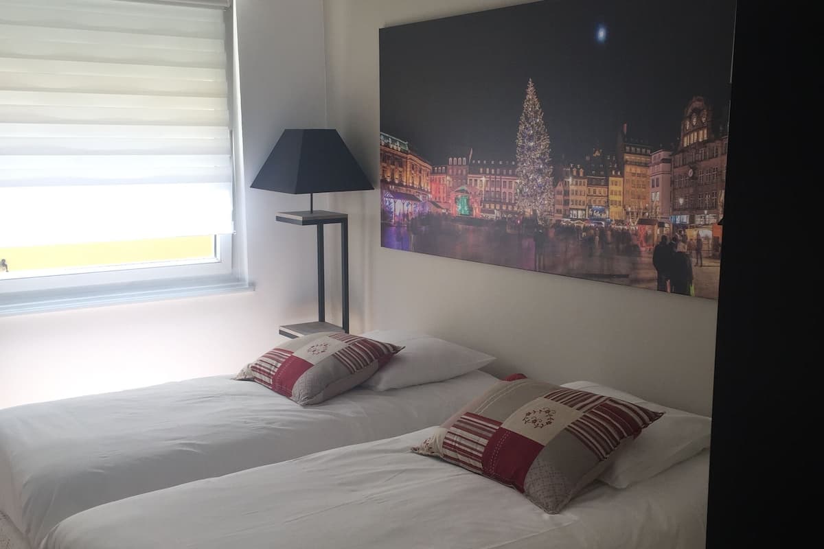 A hotel room with two beds, a large window with blinds, a floor lamp, and a cityscape painting on the wall.