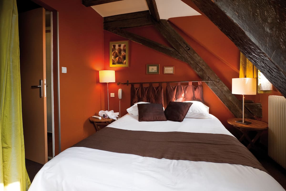 A bedroom with a large bed, wooden beams, a nightstand with a lamp, and a window with curtains.