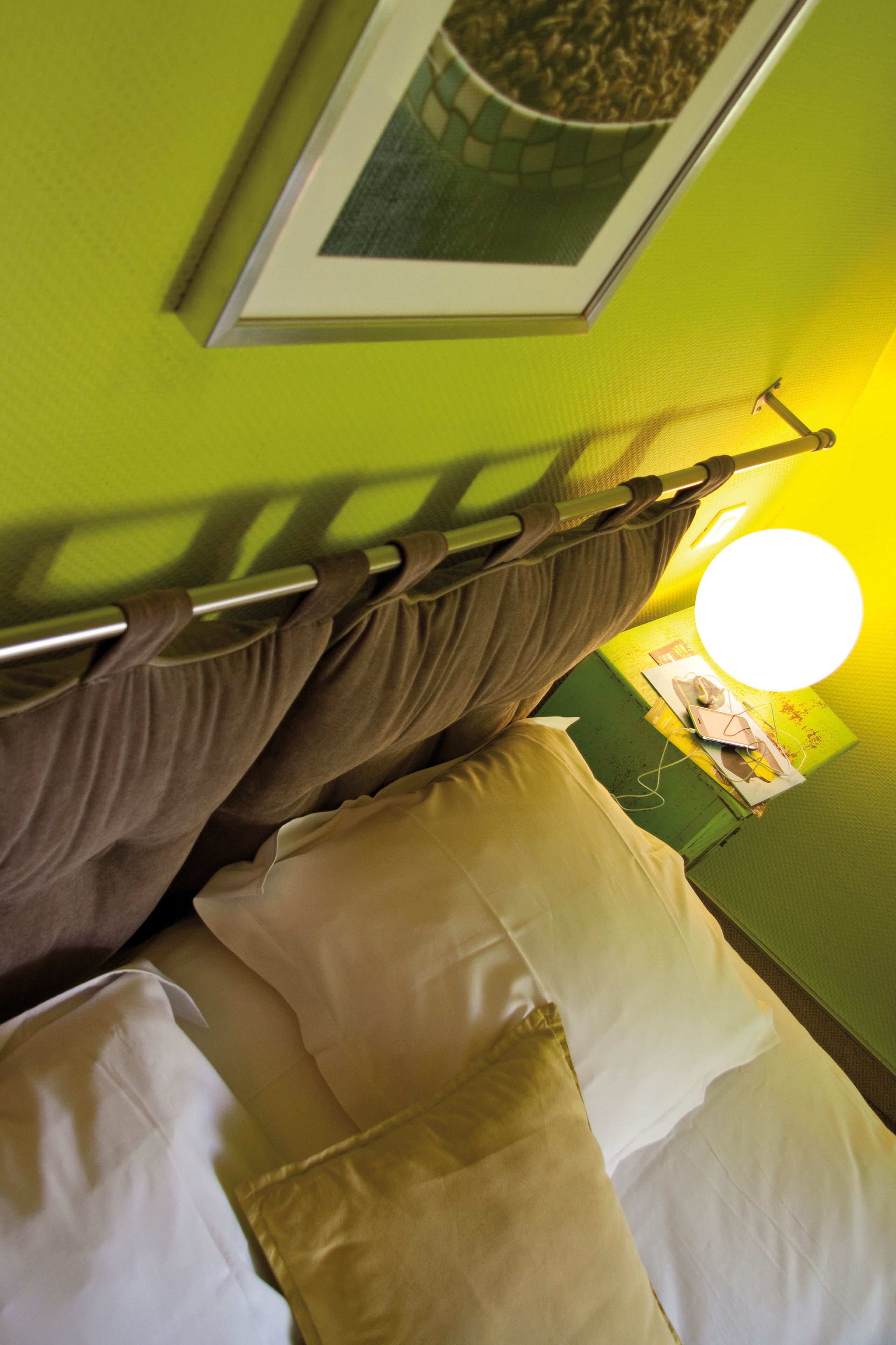 A bed with white and beige pillows, a brown headboard, and a green wall with a framed picture above it.