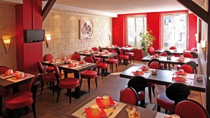 A restaurant with red chairs and tables, a TV mounted on the wall, and a large window with a view of buildings.