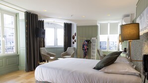 A spacious bedroom with a large bed, a sitting area with chairs, a wardrobe, and a view of the city through the window.