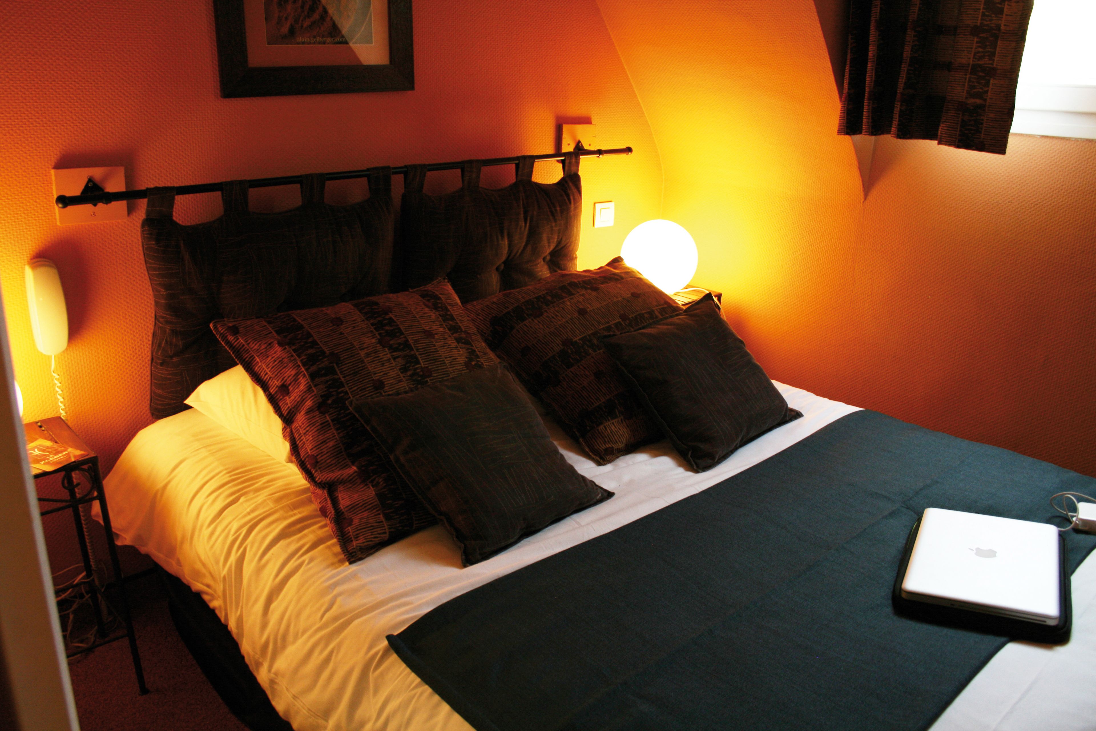 A neatly made bed with a laptop on it, a bedside lamp, and a wall-mounted picture.