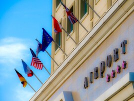 Un edificio con el letrero "AIRPORT HOTEL" y varias banderas, entre ellas la estadounidense, la de la Unión Europea y otras.