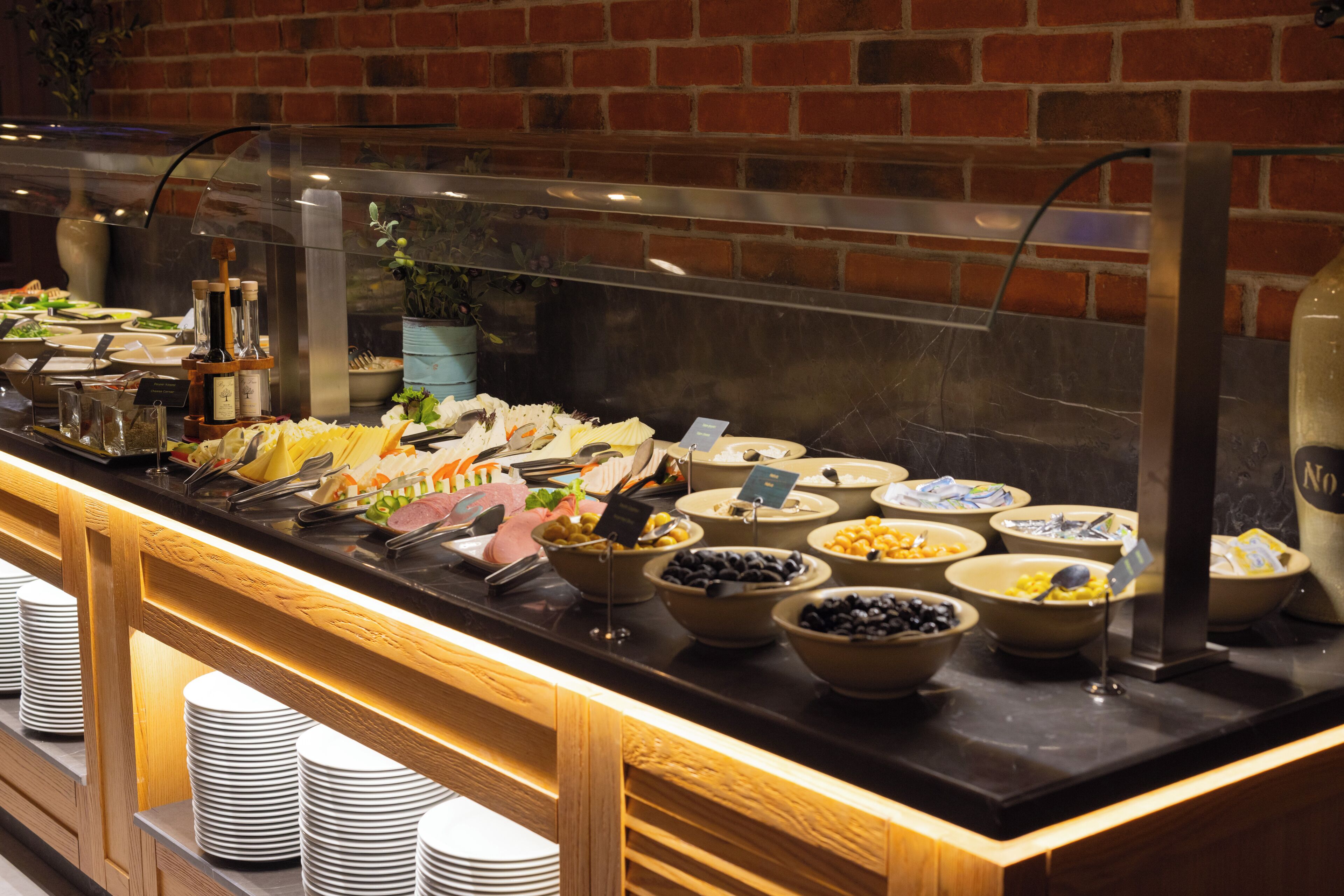 A buffet with various food items and condiments, including bowls of fruits, vegetables, and meats, with a brick wall in the background.
