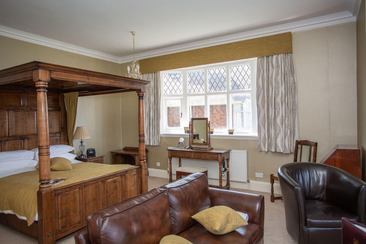 A four-poster bed, a leather sofa, a wooden desk, and a chair in a room with a large window and a radiator.