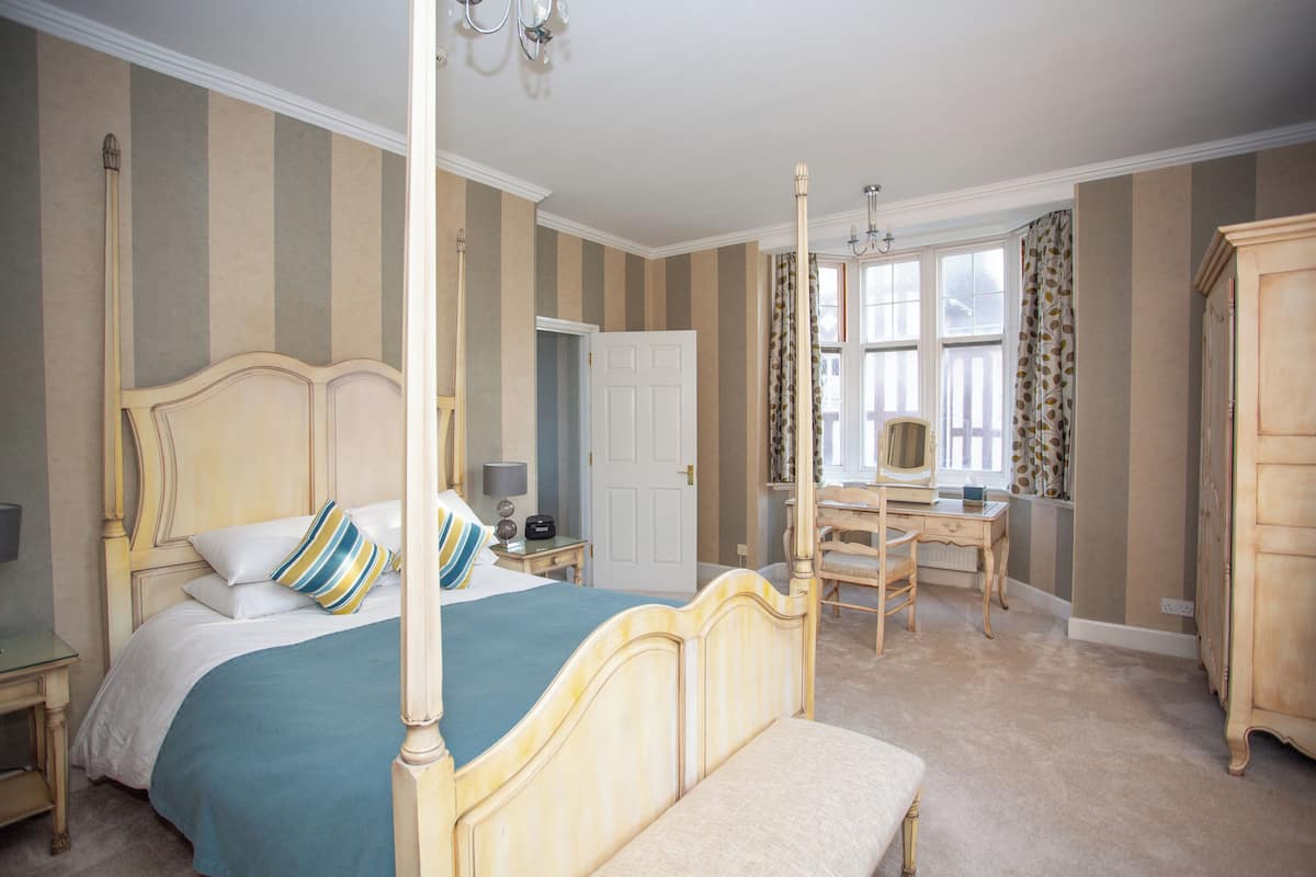 A four-poster bed with a bench, a desk, and a wardrobe in a room with striped wallpaper and a large window.