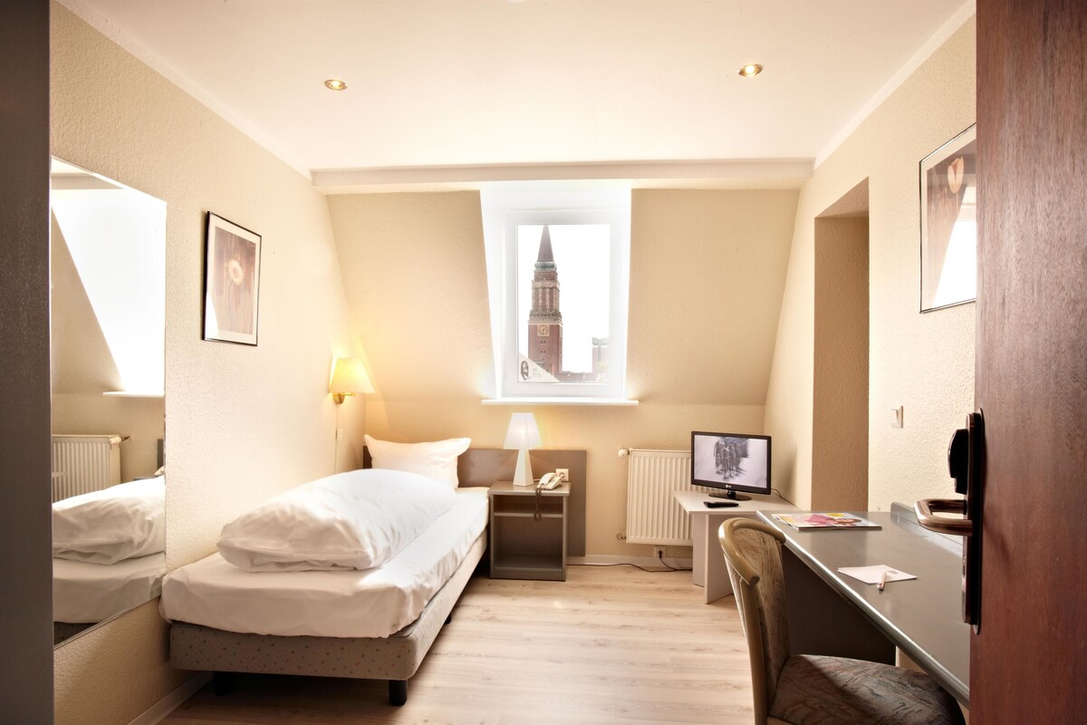 Ein Hotelzimmer mit einem Bett, einem Schreibtisch mit Computer, einem Sessel, einer Lampe, einem Nachttisch und einem Fenster mit Blick auf einen Turm.