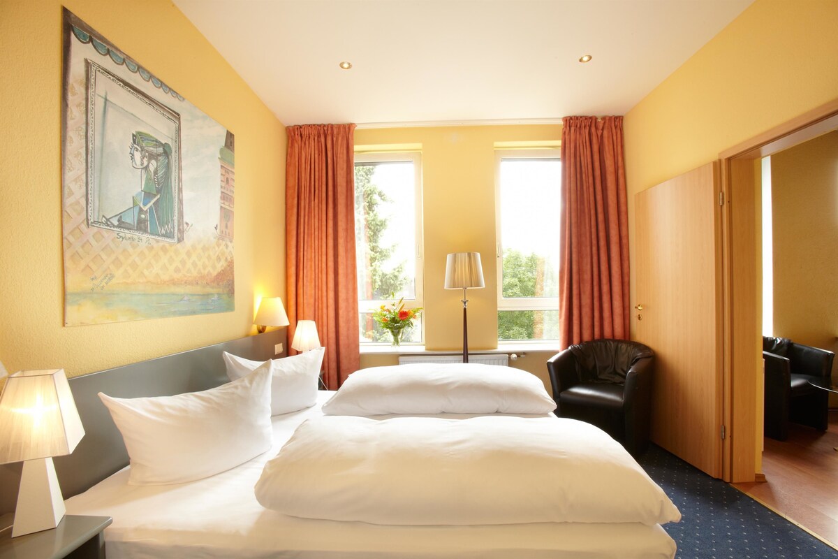 Ein Hotelzimmer mit einem großen Bett, zwei Nachttischen mit Lampen, einem Sessel, einem Schreibtisch und einem Vorhangfenster.